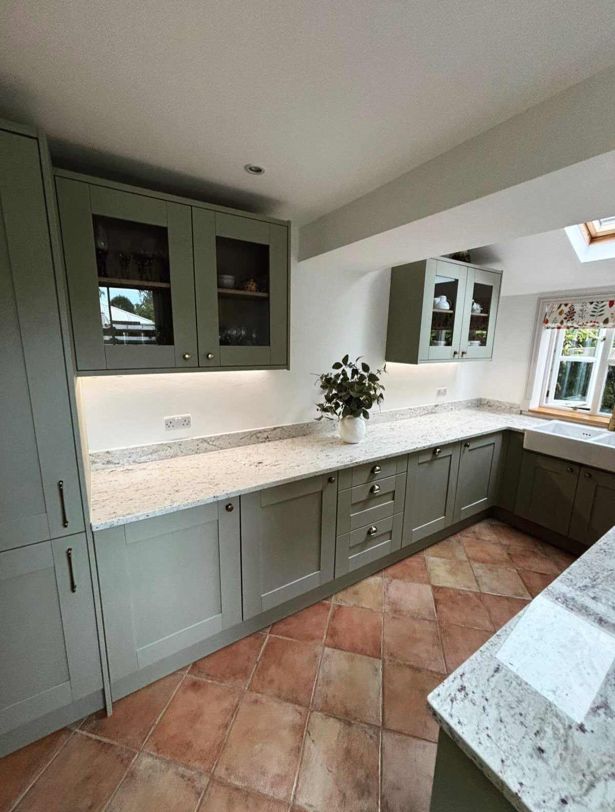 Sage green Shaker cabinetry with stone countertops and terracotta tile flooring in a kitchen.