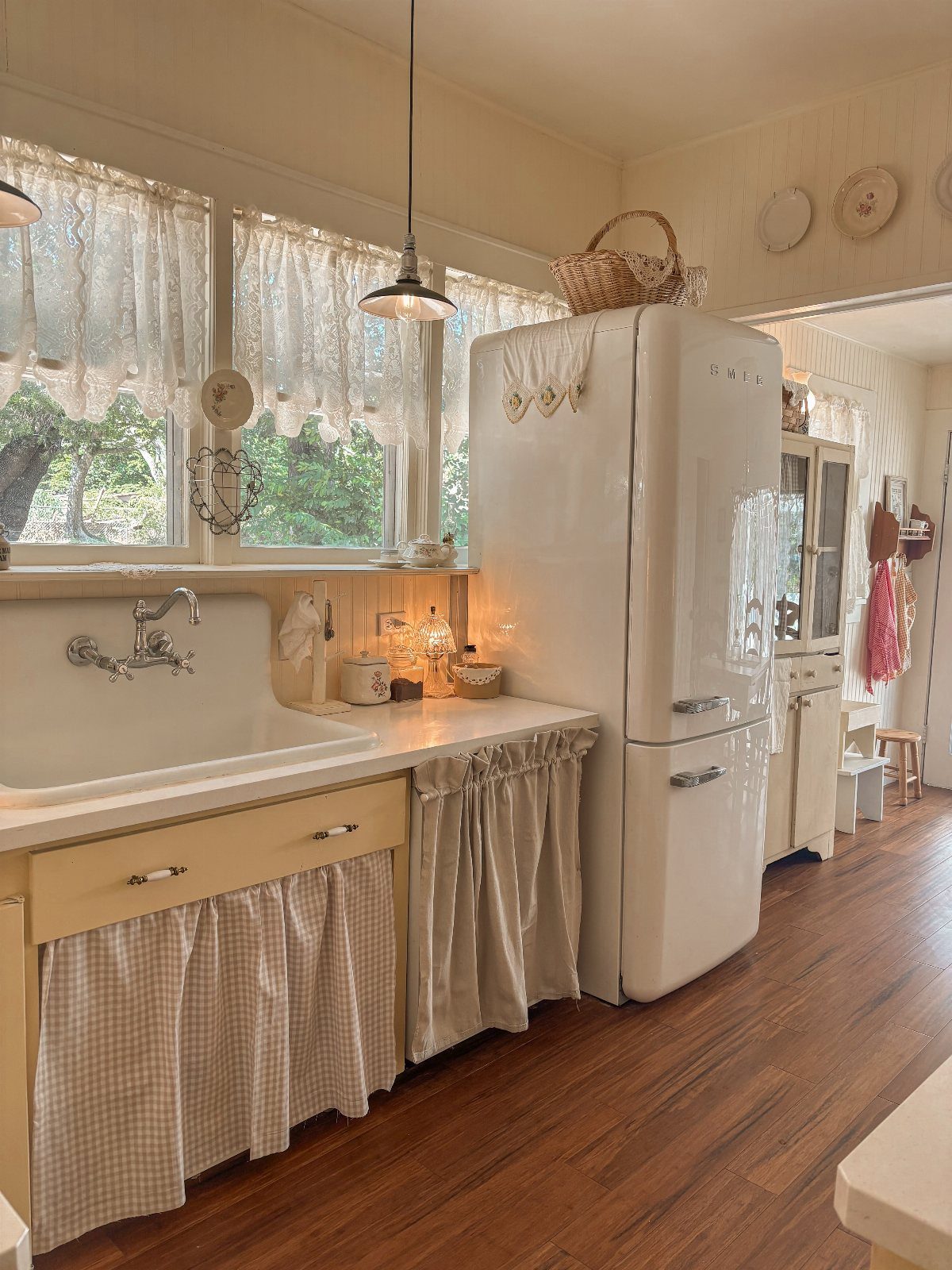Cream colored kitchen with vintage refrigerator, wood floors, and lace window curtains.
