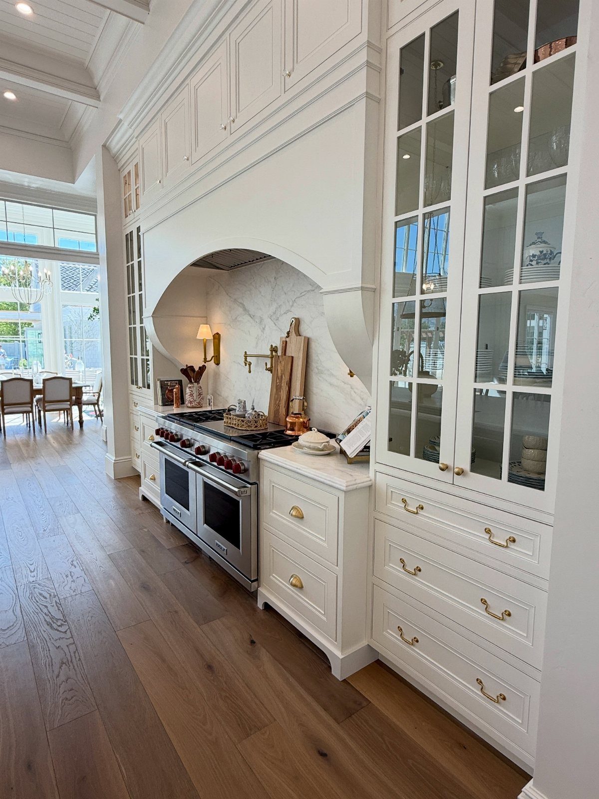 White traditional kitchen with marble range hood, gold hardware, and light wood flooring.