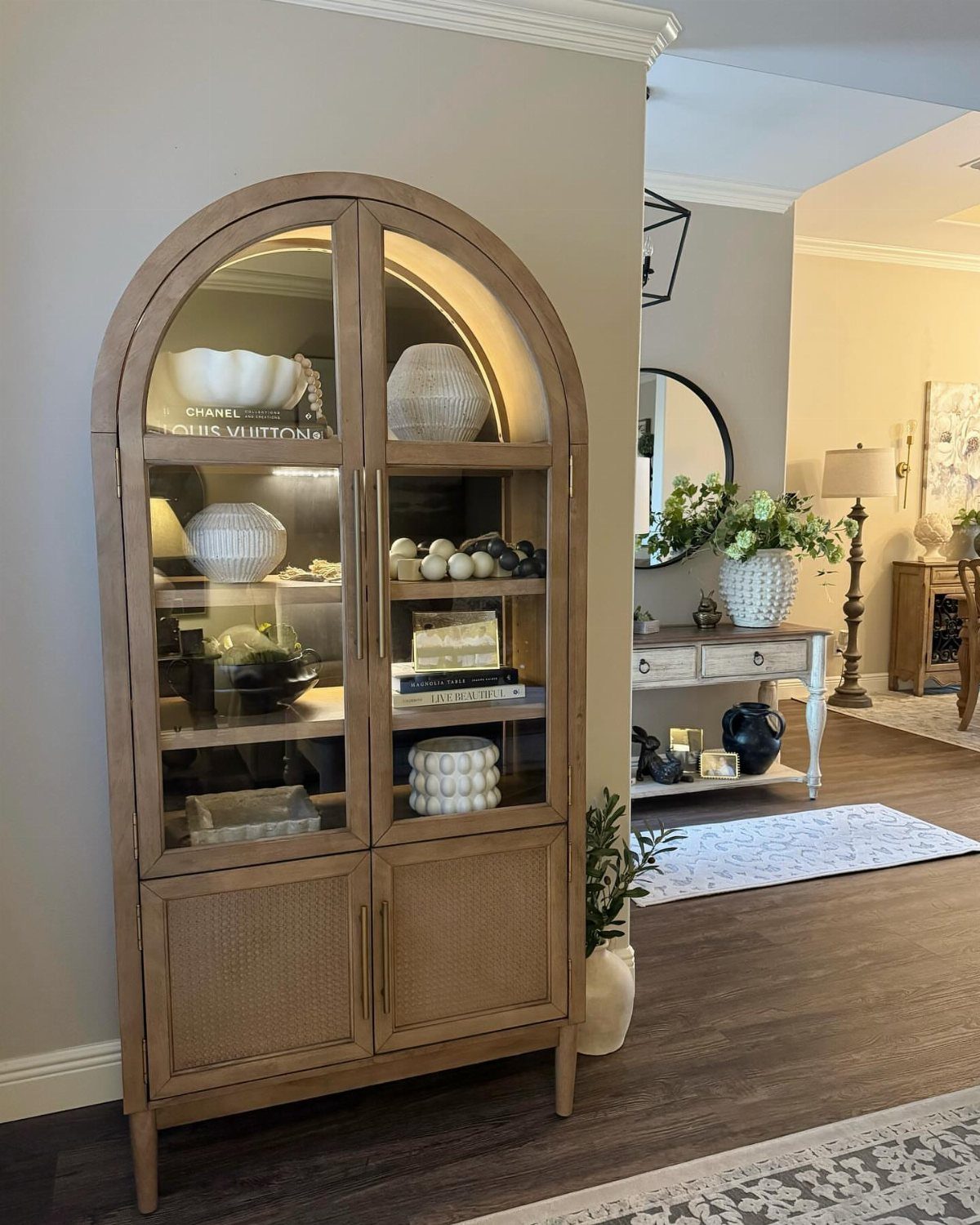 Arched wooden storage console with glass display and cane doors against a grey wall.