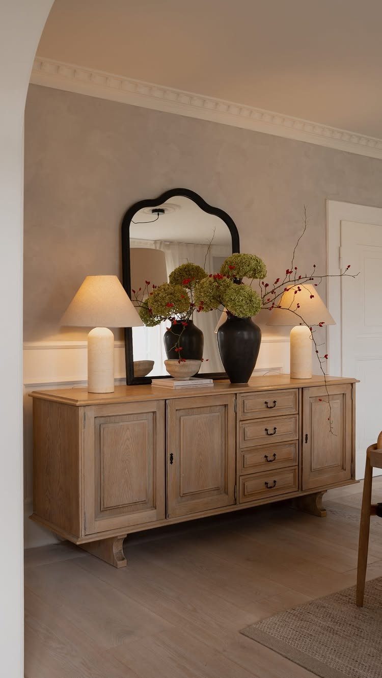 Wooden console table with two cream table lamps, a black mirror, and green flowers.