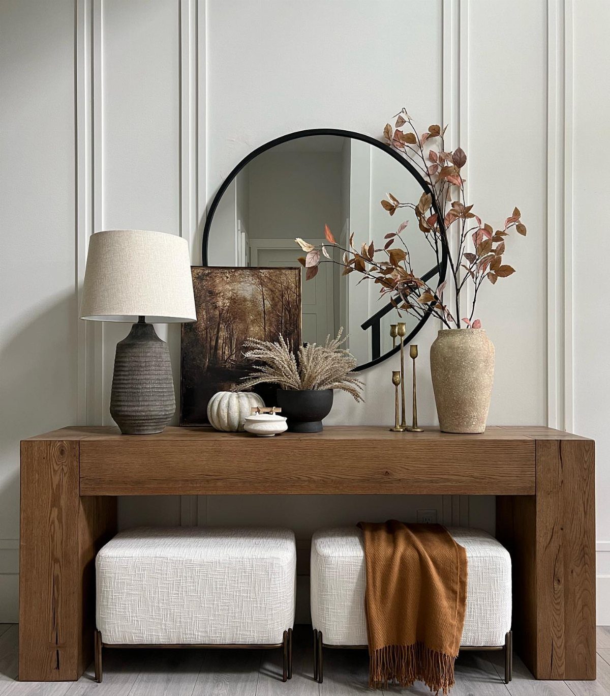Chunky wood console table with a round mirror, lamp, and white poufs underneath.