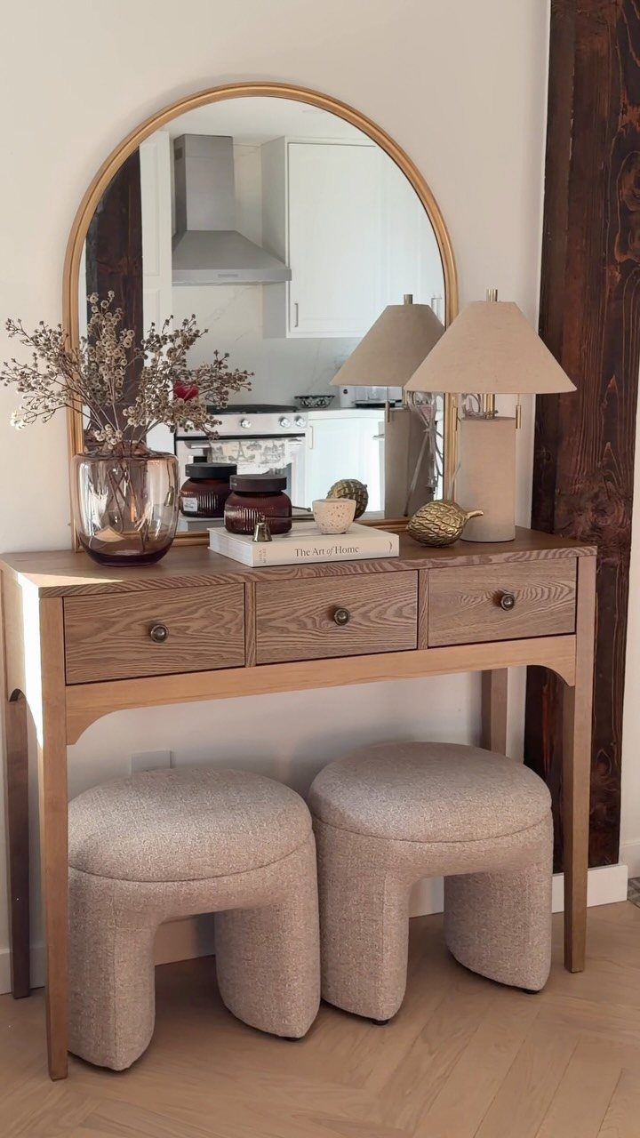 Light wood console table with drawers and poufs under an arched mirror.