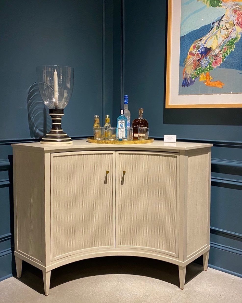 Curved light-colored corner cabinet with liquor bottles and a candle holder on top.