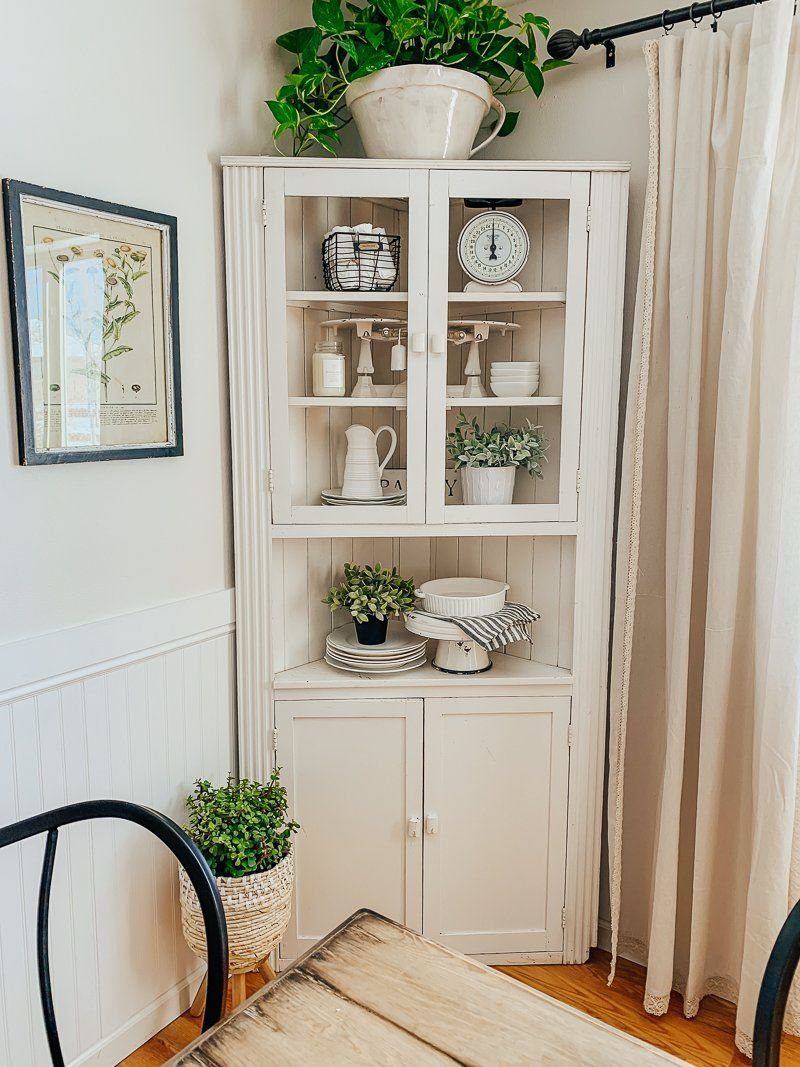 White corner cabinet with glass doors displaying white ceramics and a clock.
