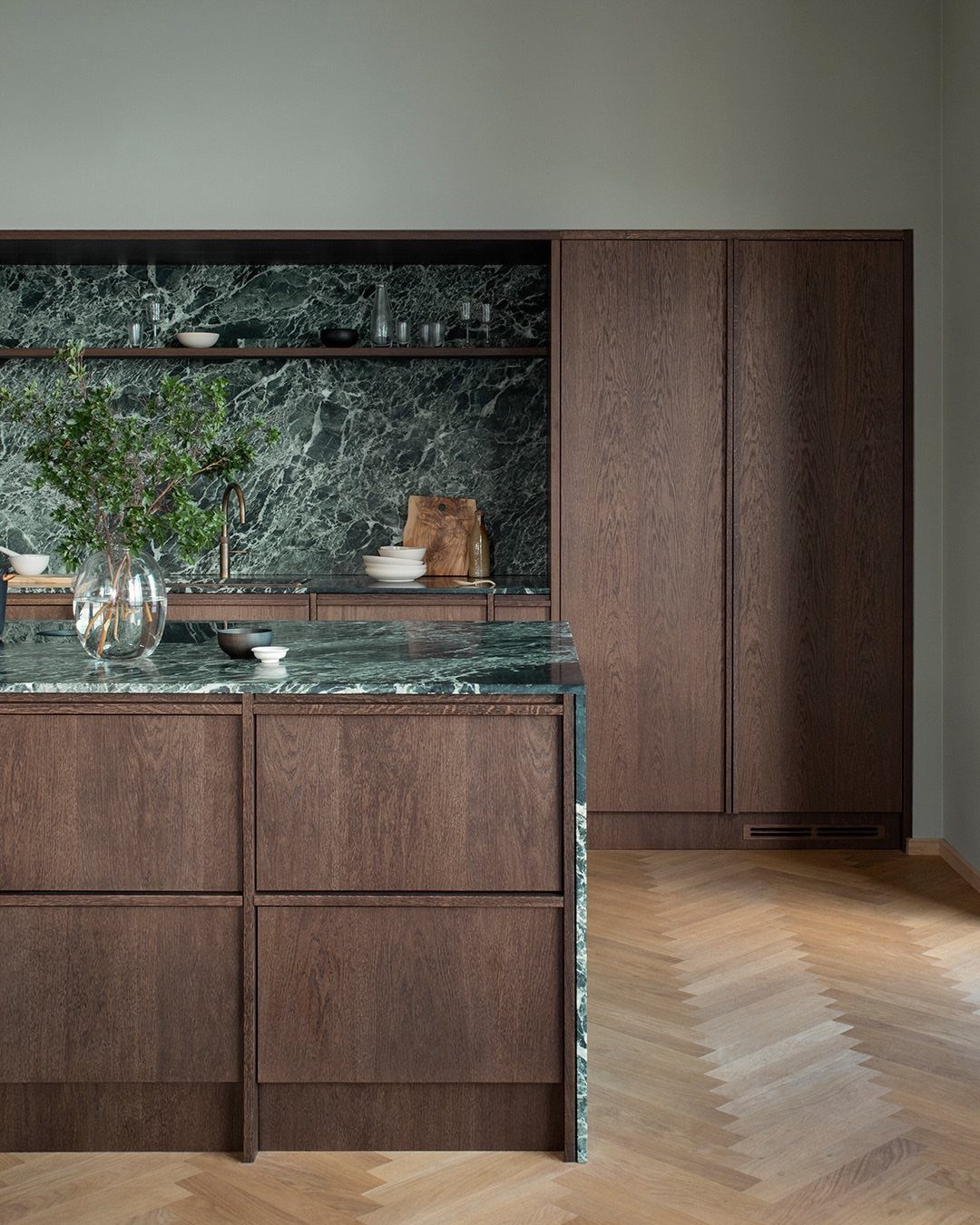 Dark brown wood kitchen cabinets with green marble countertops and herringbone wood floors.