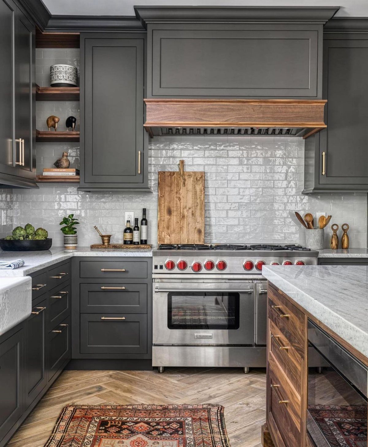 Charcoal gray kitchen cabinets with gold hardware, marble countertops, and a wood range hood.