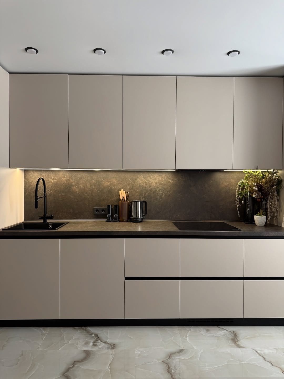Modern kitchen with taupe cabinets, dark backsplash, black countertops, and white marble flooring.