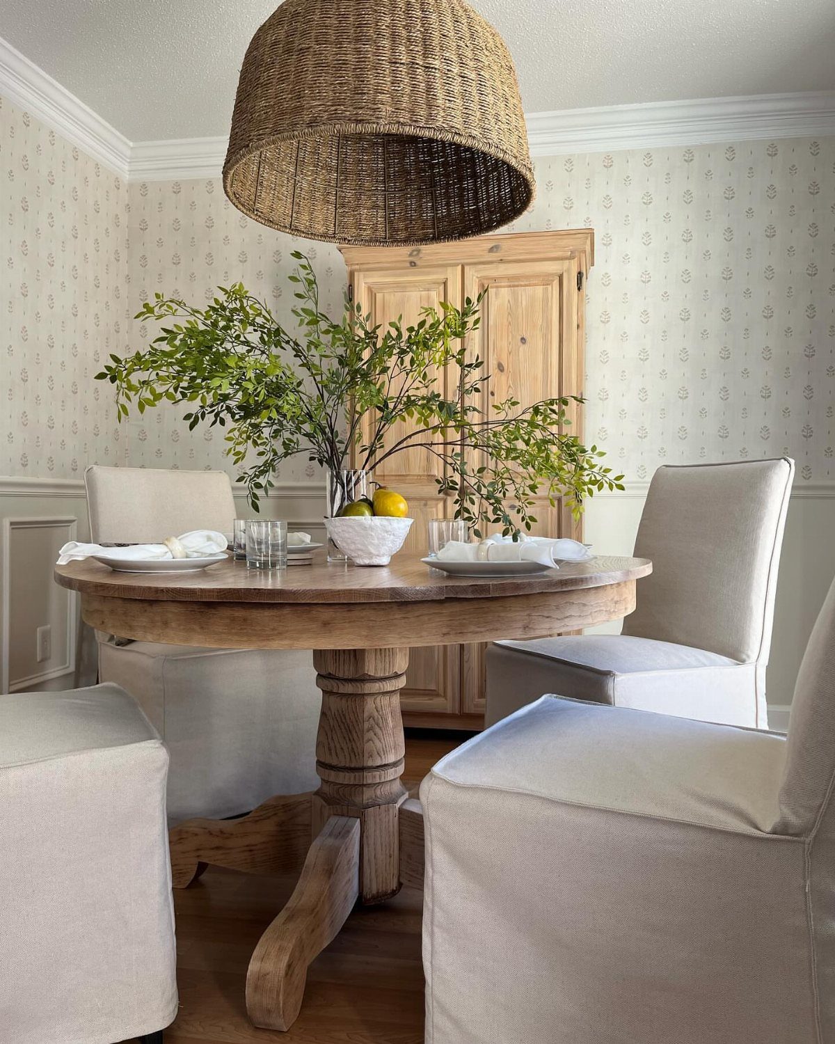 Round wooden pedestal table with cream upholstered chairs under a large wicker pendant light.
