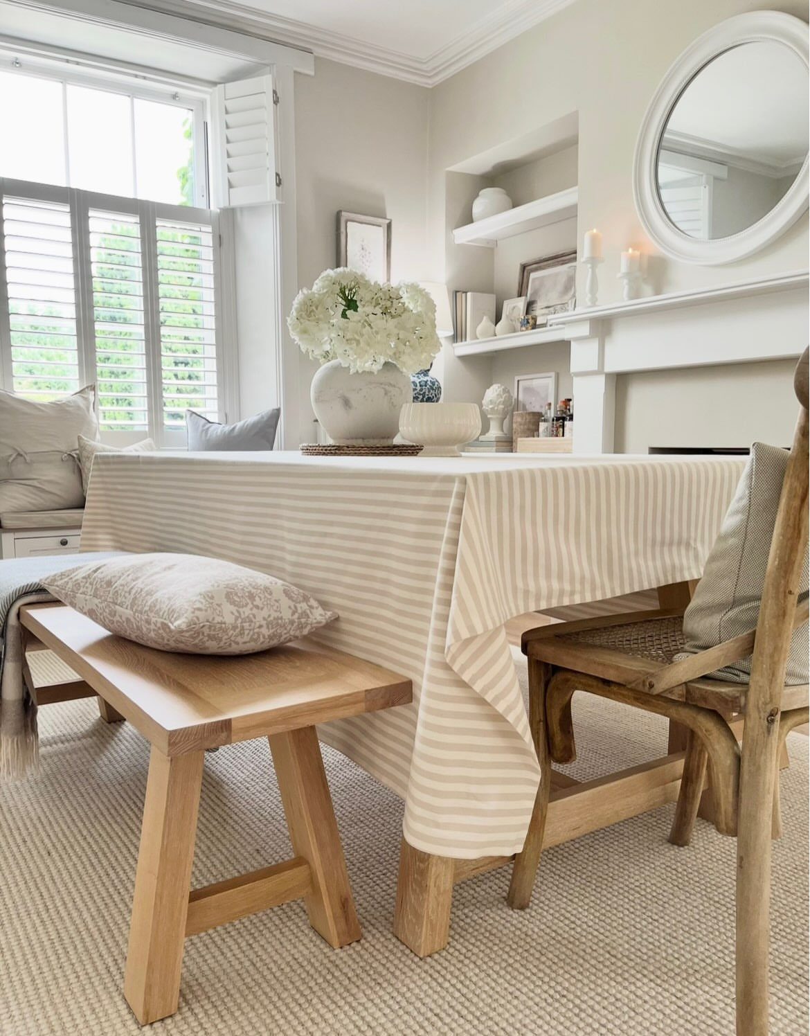 Wooden dining table with striped tablecloth, bench, and woven chairs in a bright room.