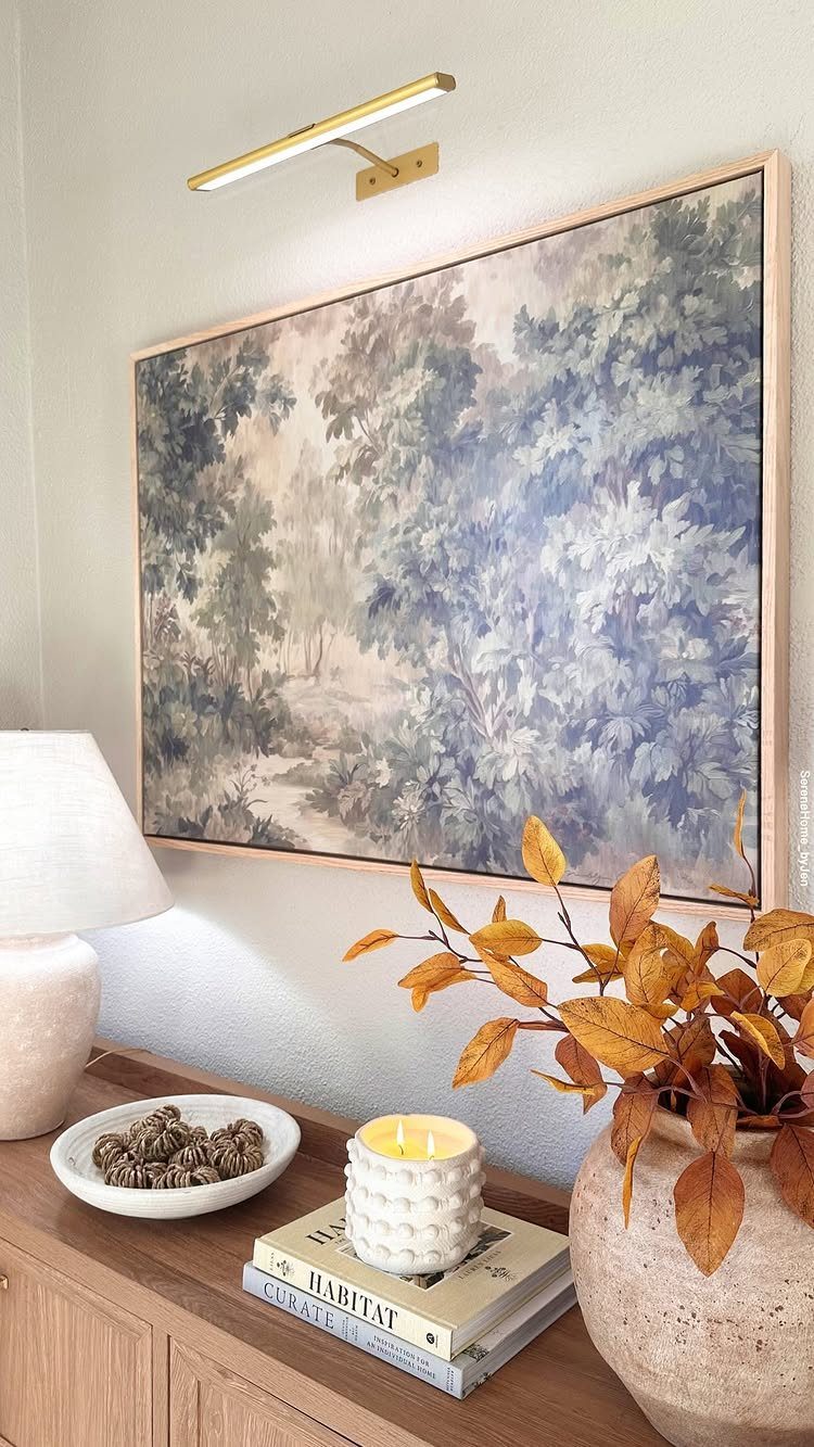 Wooden sideboard with books, a candle, and a vase of autumn leaves below a painting.