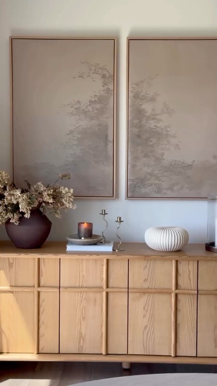 Light wood sideboard decorated with a vase, candle, and bowl under two large paintings.