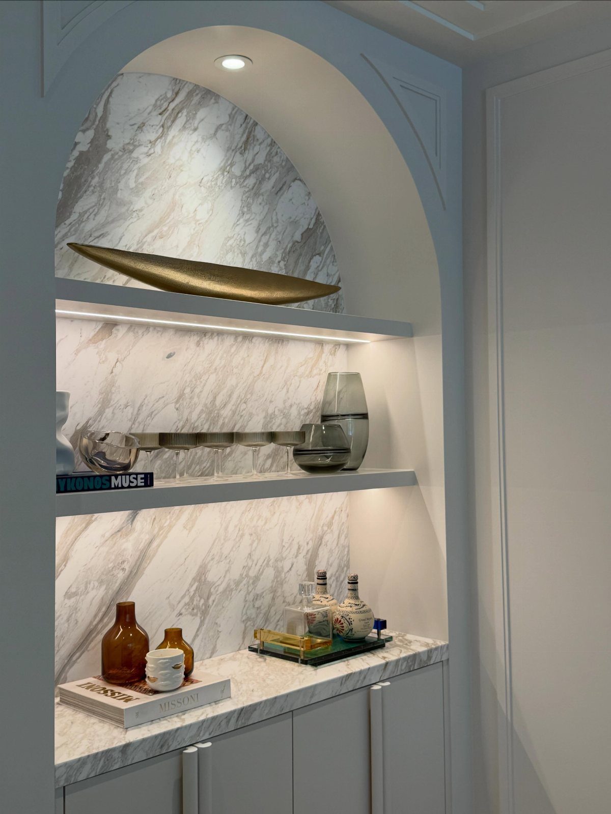 White built-in sideboard with marble surfaces and floating display shelves in an arched alcove.