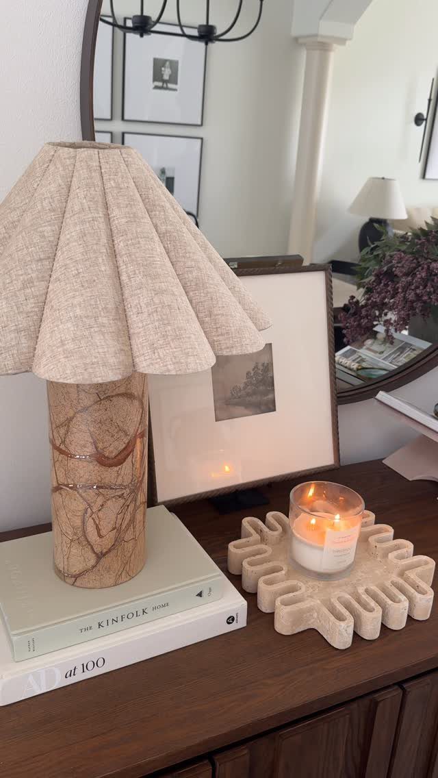 Lit candle on a wavy cream tray atop a wooden sideboard with a lamp.