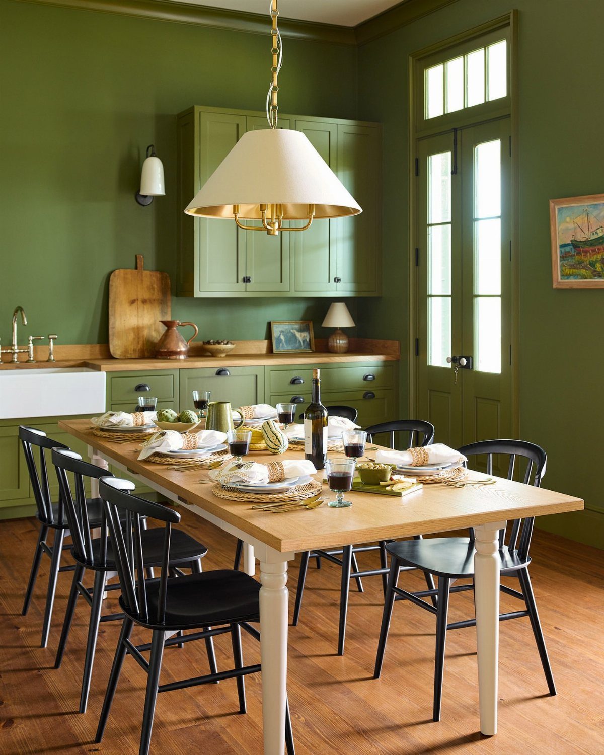 Light wood dining table with black chairs in a green dining room setting.