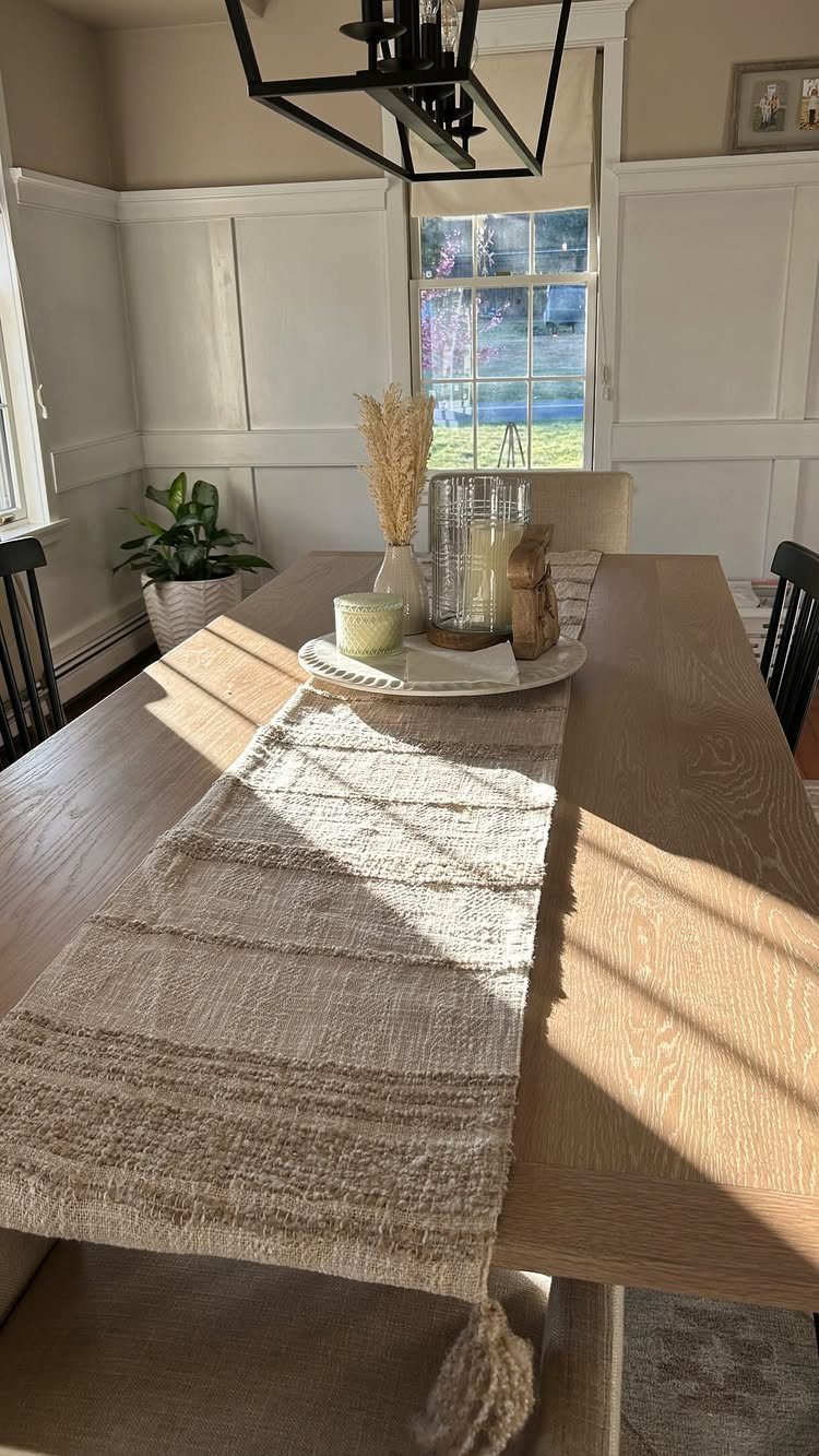 Light oak dining table with a textured runner and decorative candles in a bright room.