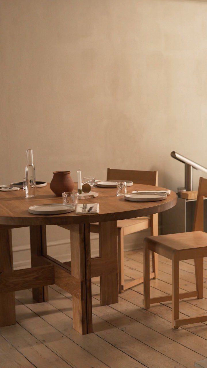 Round wooden dining table with trestle base and matching chairs in a minimalist room.