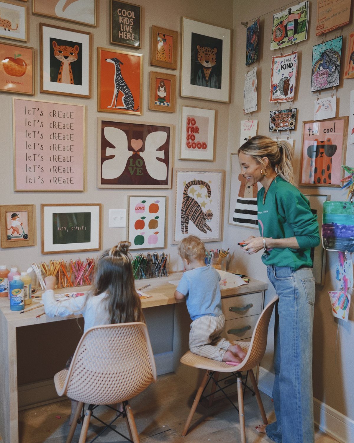 Children and a woman painting at an art table with supply jars and gallery walls.