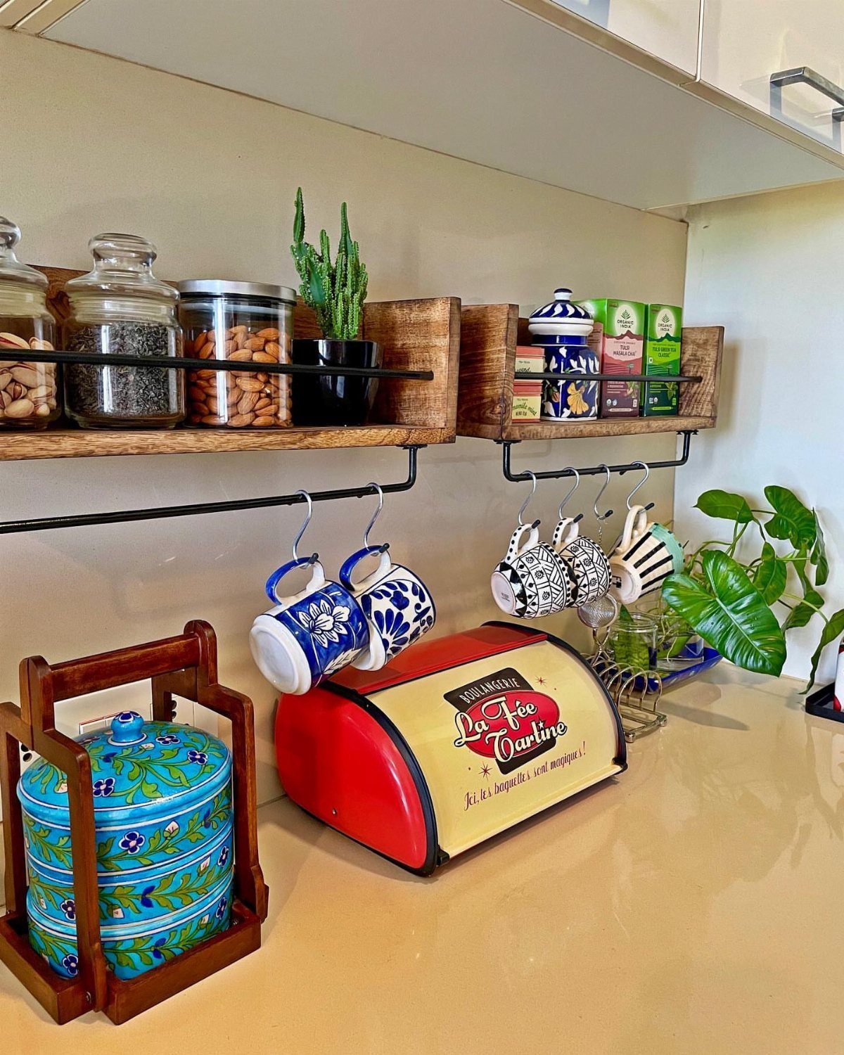 Kitchen counter with wooden shelves, patterned mugs, red bread box, and blue tiffin carrier.