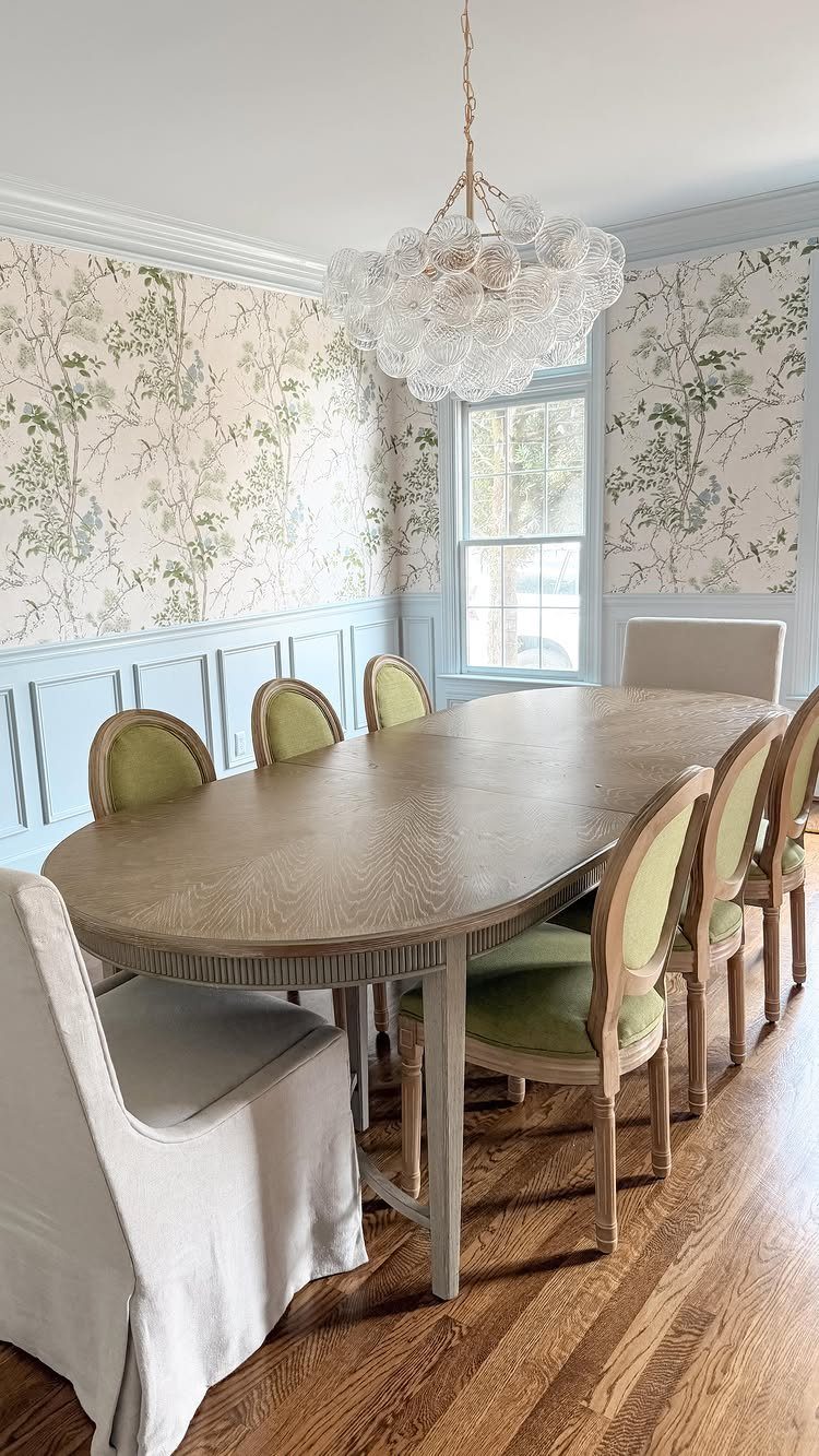 Oval wooden dining table with green upholstered chairs in a floral wallpapered room.