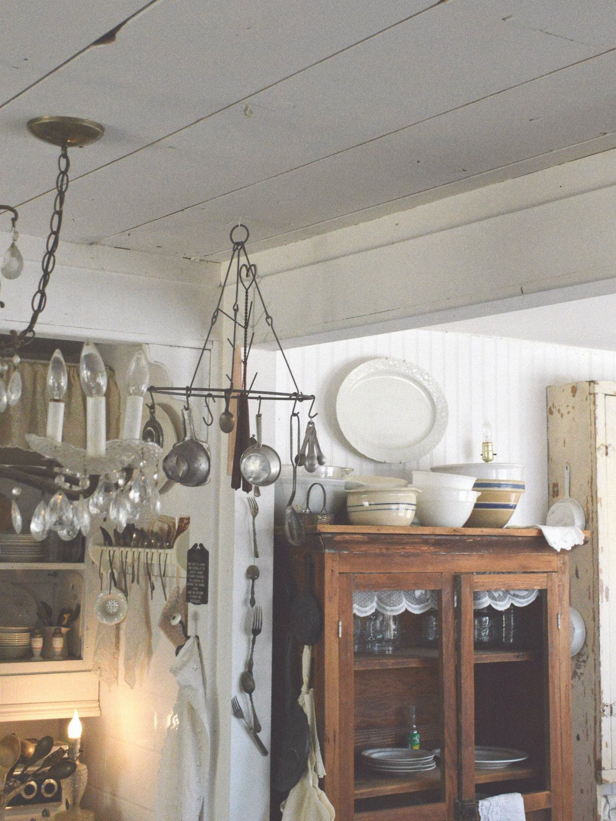 Metal hanging pot rack with cookware in a rustic white kitchen with wooden hutch.