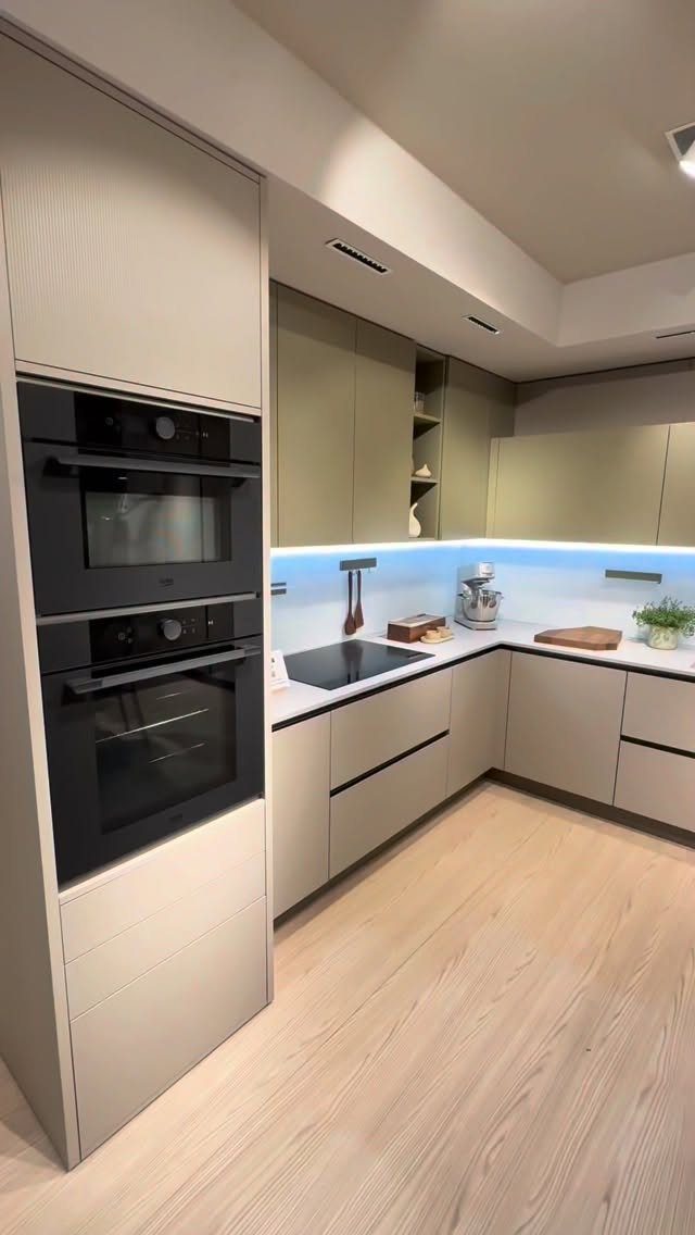 Modern kitchen with light beige minimalist cabinets, integrated black ovens, and light wood floors.
