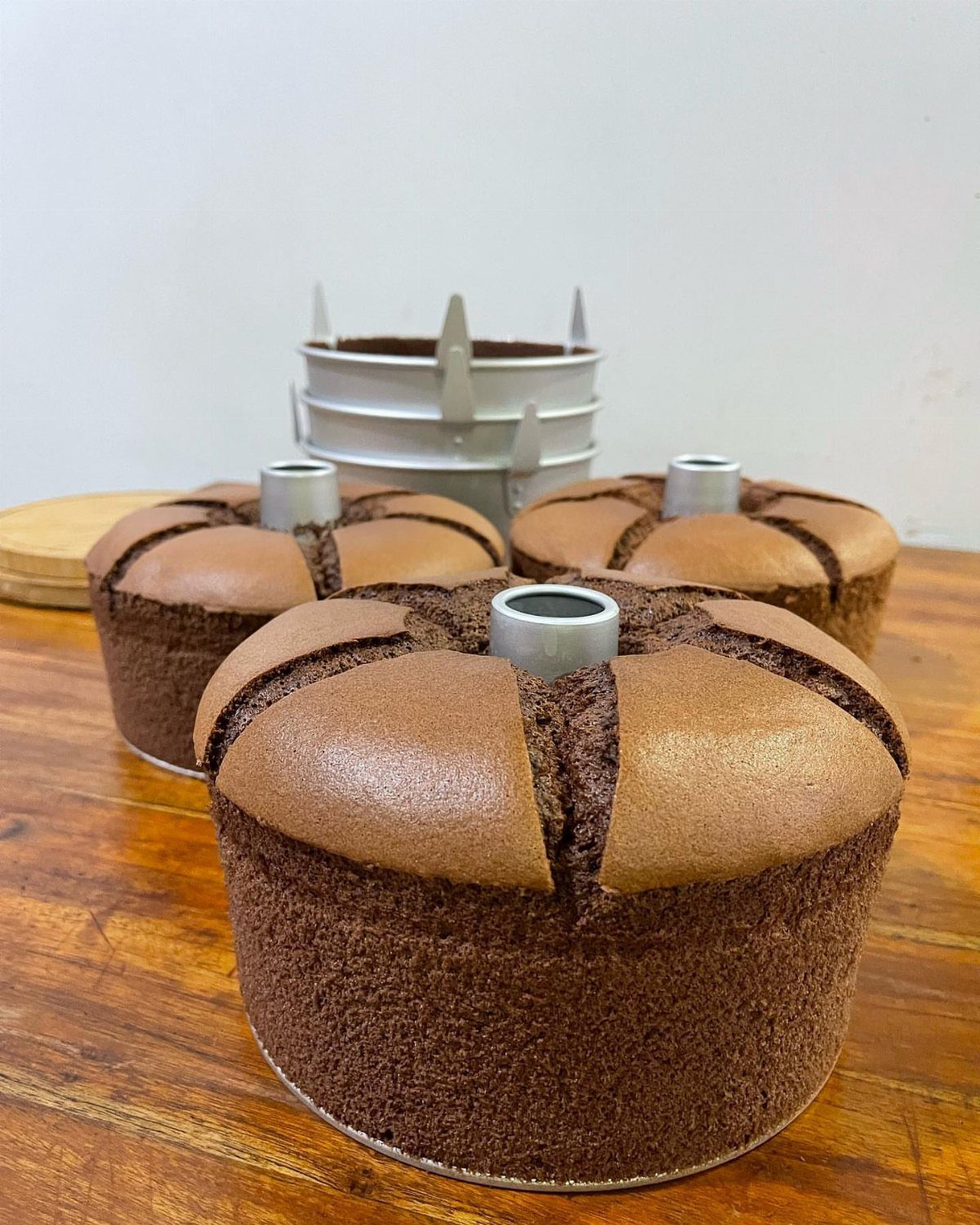 Three chocolate chiffon cakes in tall silver pans on a wooden table.