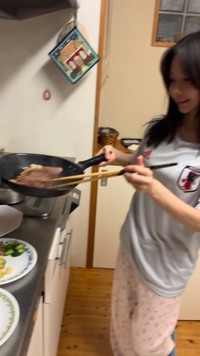 Woman using long wooden cooking chopsticks to fry meat in a black pan.