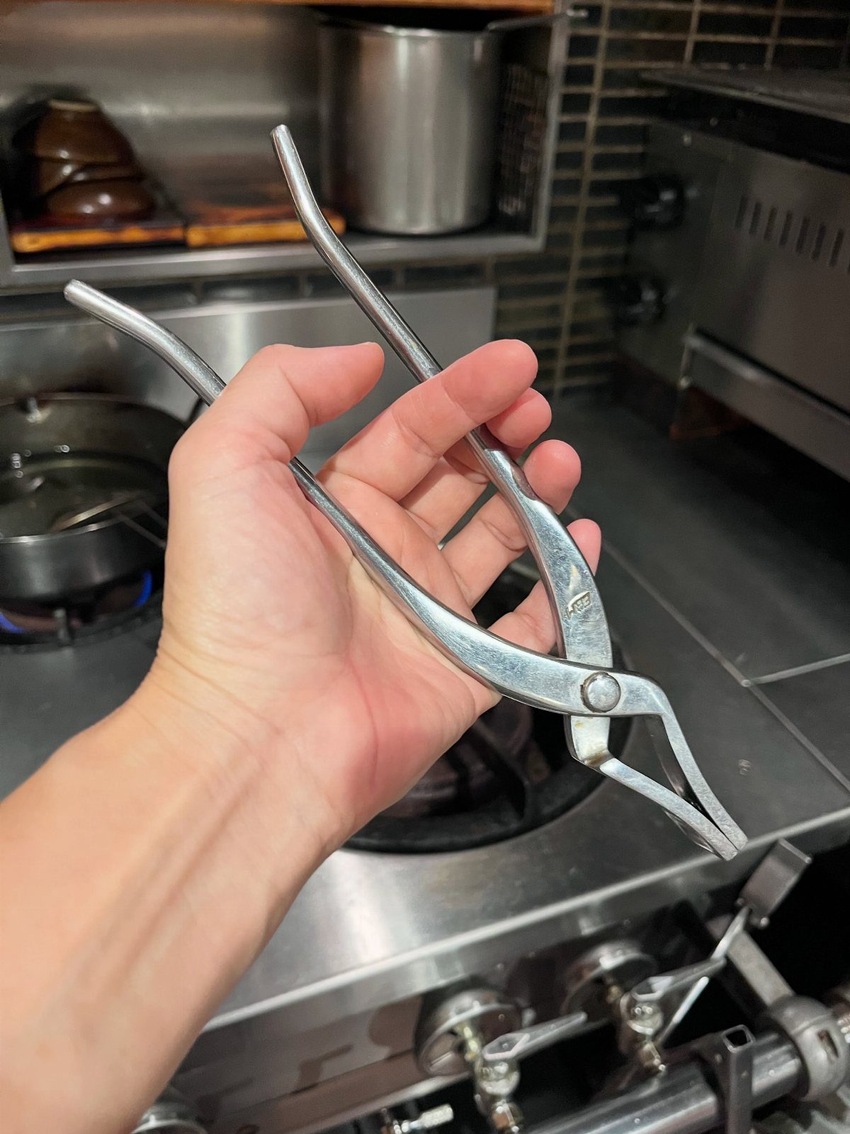 Silver metal cooking tongs held by a hand in a professional kitchen.