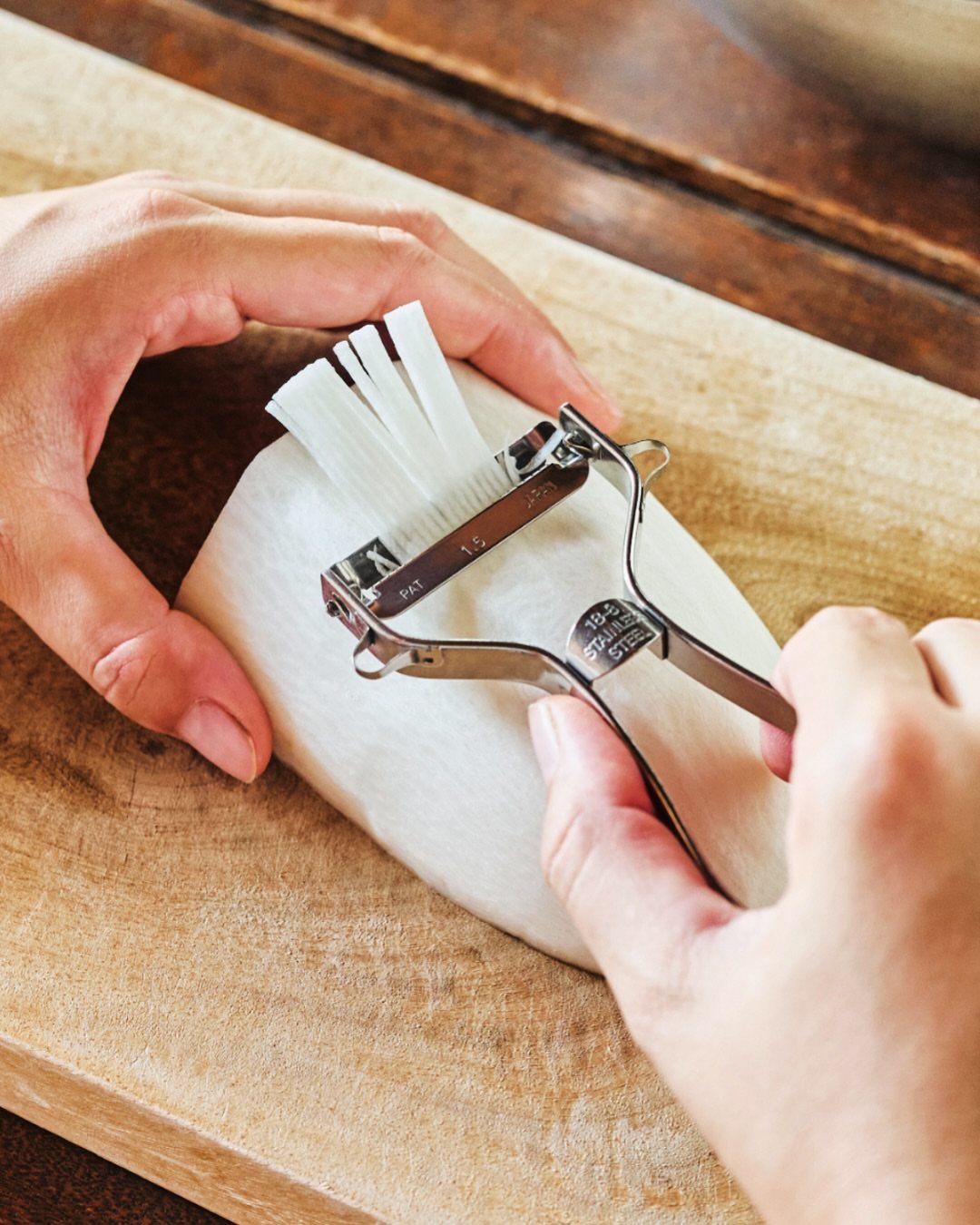 Hands using a stainless steel slicer on a white daikon radish on wood.