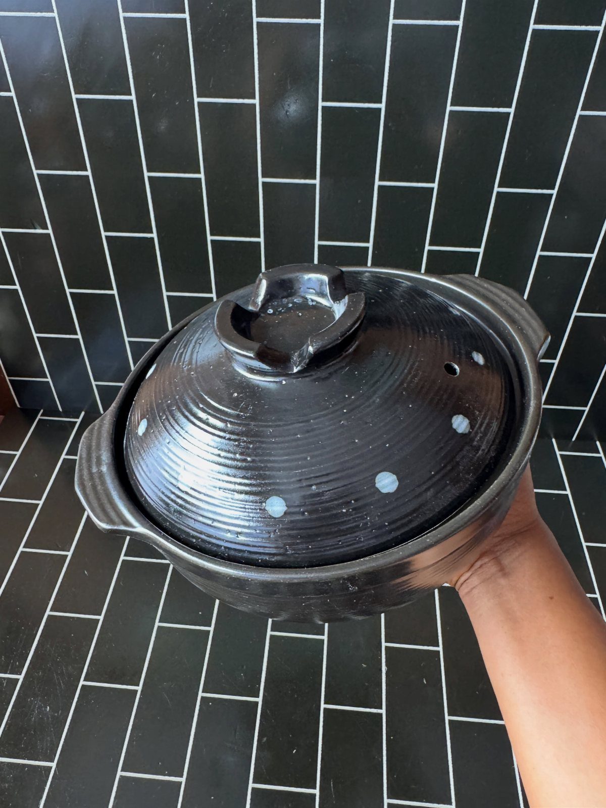 Black ceramic Japanese donabe pot with lid held against black tiled wall.