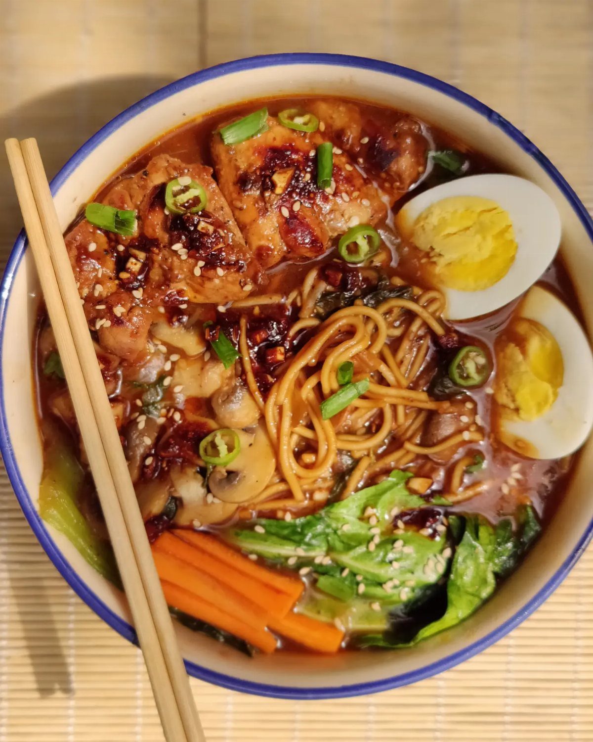 Ramen bowl with noodles, eggs, and vegetables on a bamboo mat with chopsticks.