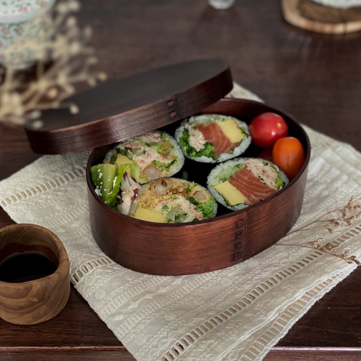 Round wooden Magewappa bento box filled with colorful sushi rolls and cherry tomatoes.