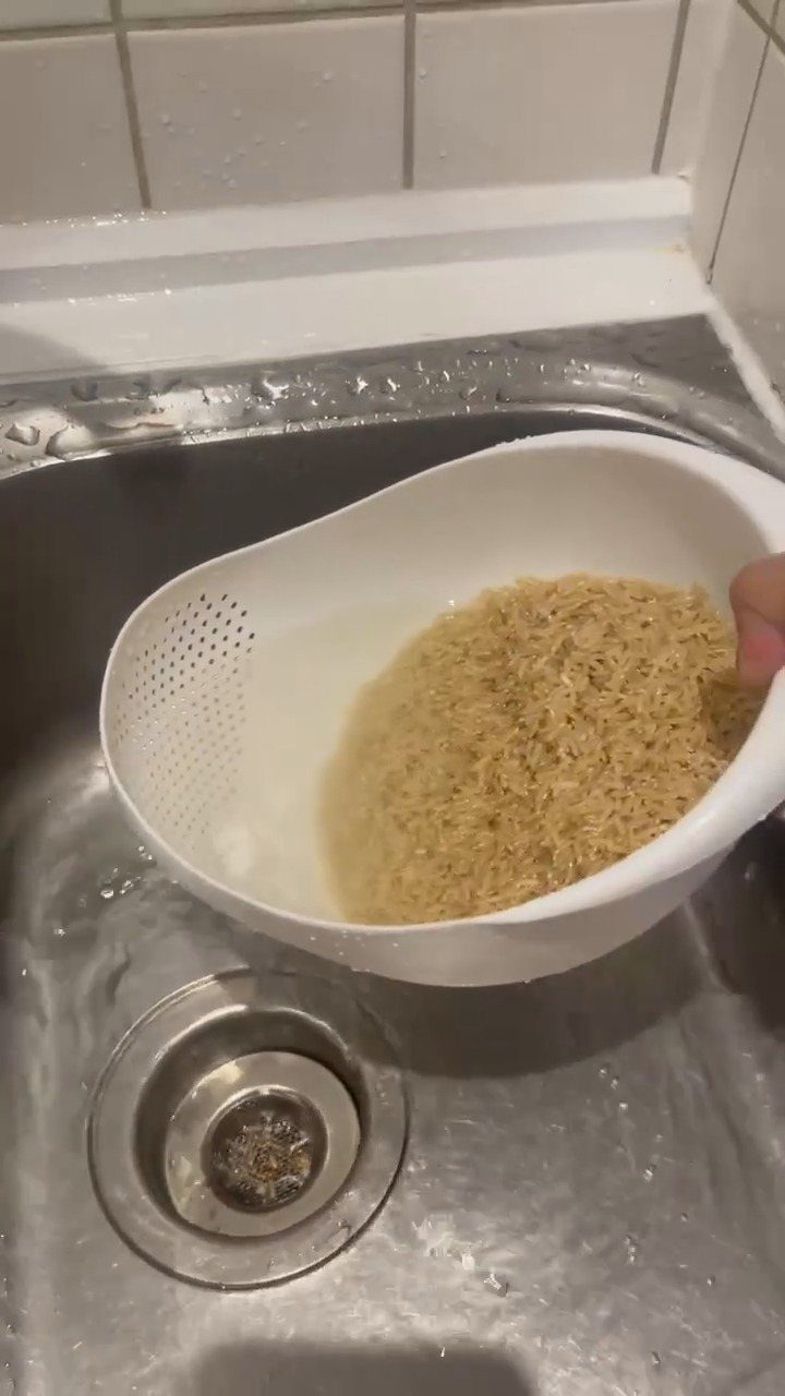 White plastic rice washing bowl with brown rice held over a metal sink.