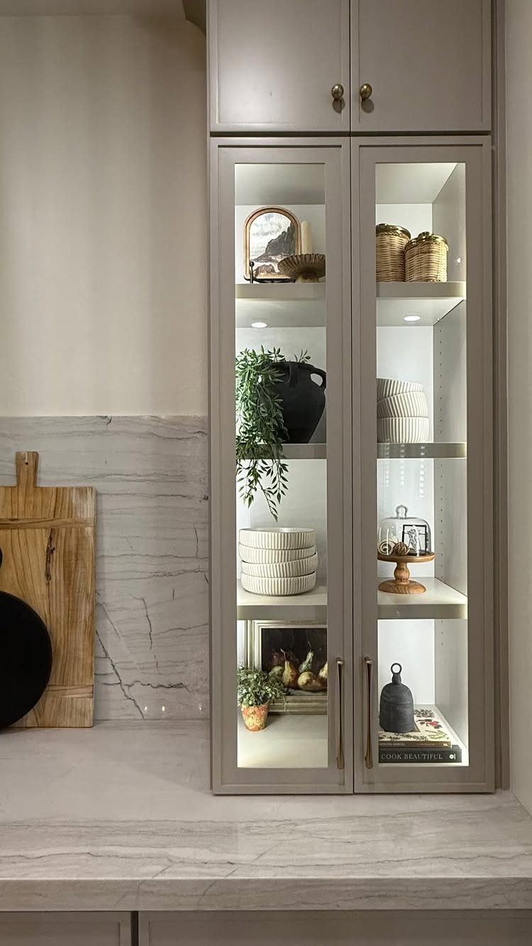 Gray kitchen cabinet with glass doors and interior lighting on white marble countertop.