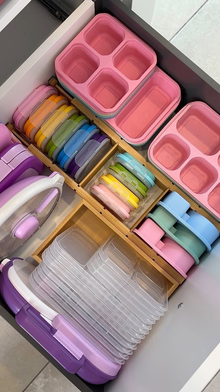 Colorful plastic food containers organized within wooden dividers inside a grey kitchen drawer.