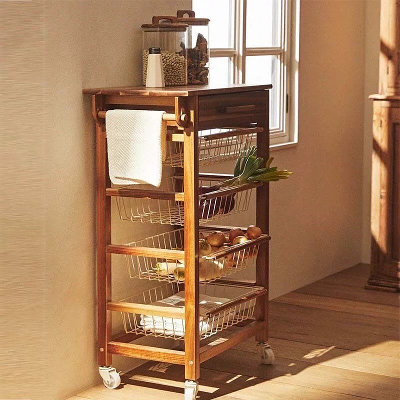 Wooden kitchen trolley with a drawer, wire baskets, and wheels in a bright room.
