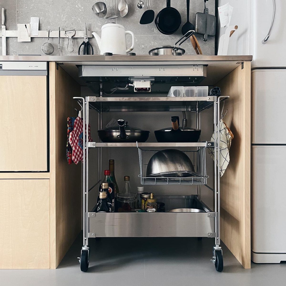 Stainless steel rolling utility cart with three tiers stored under a kitchen counter.