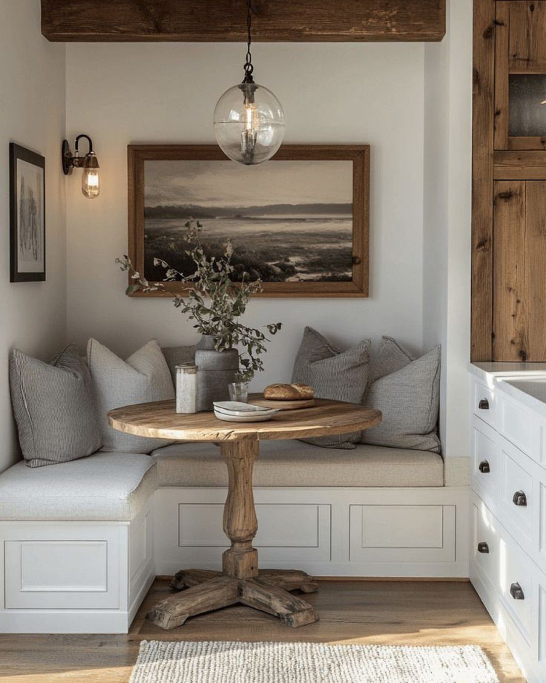 Round wooden pedestal table with white built-in banquette seating and grey pillows.