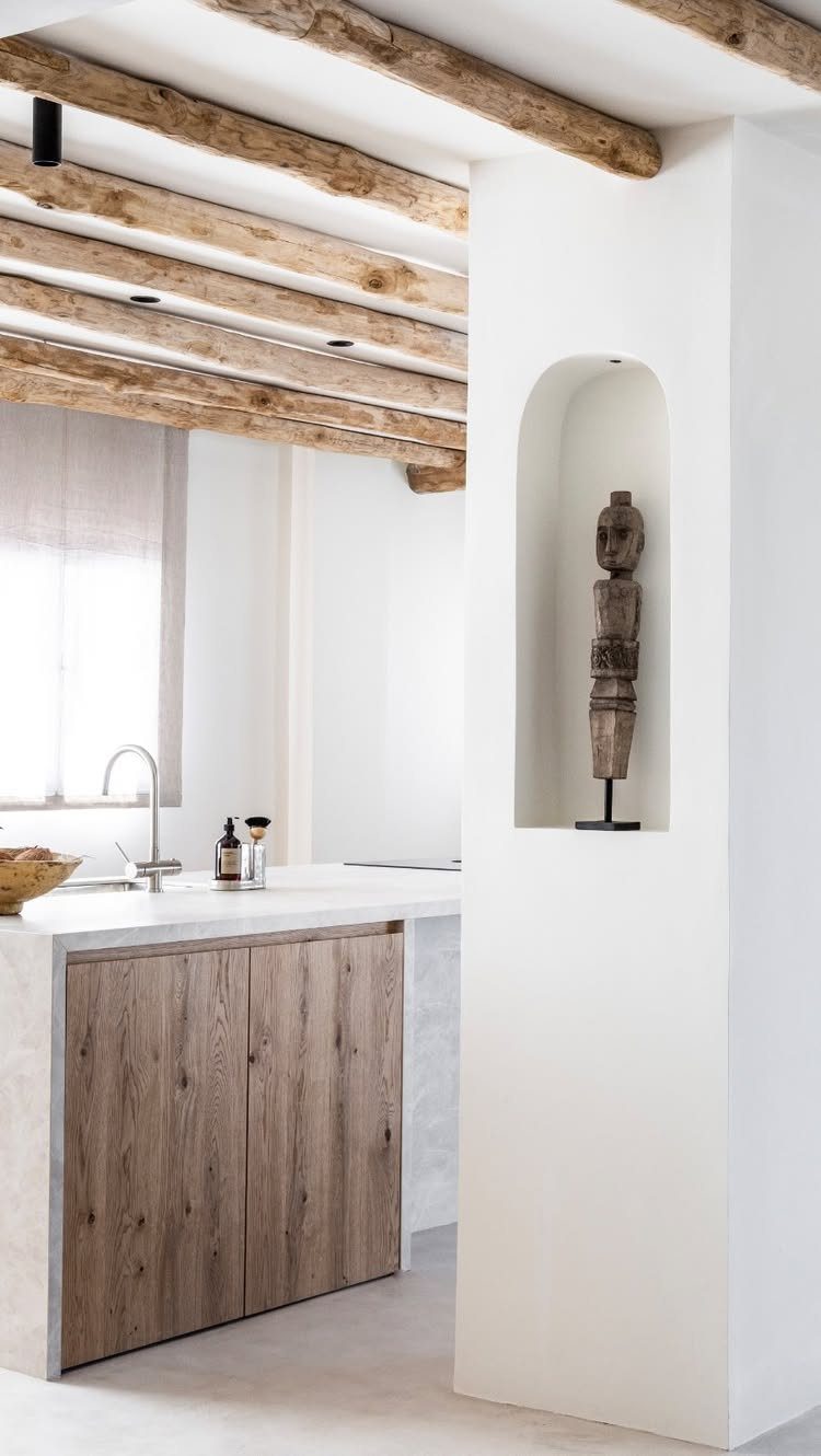 Rustic kitchen with heavy wooden ceiling beams and light wood cabinetry in white room.
