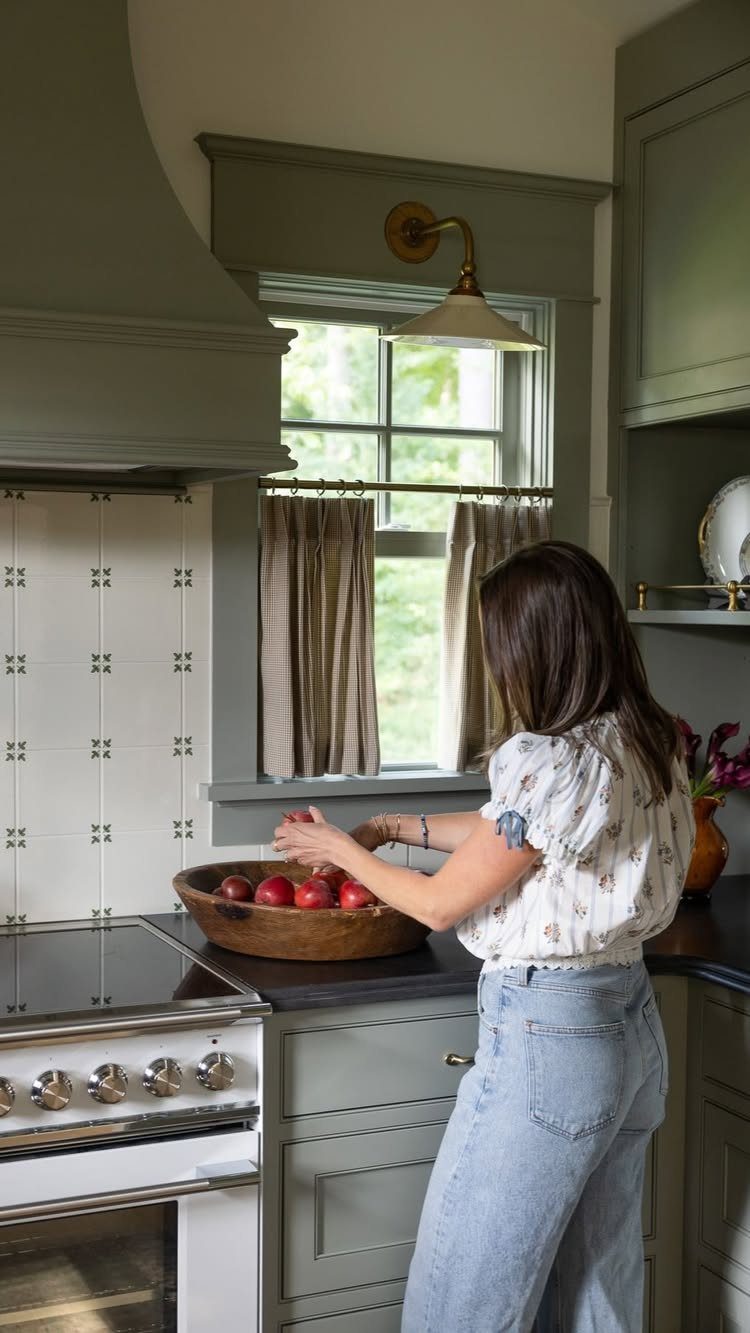 Woman preparing apples in a kitchen with sage green cabinets and white tile backsplash.