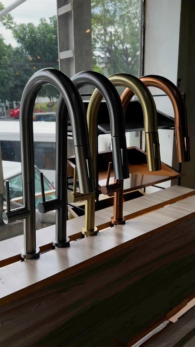 Four pull-down kitchen faucets in various metallic finishes displayed on a wooden shelf.