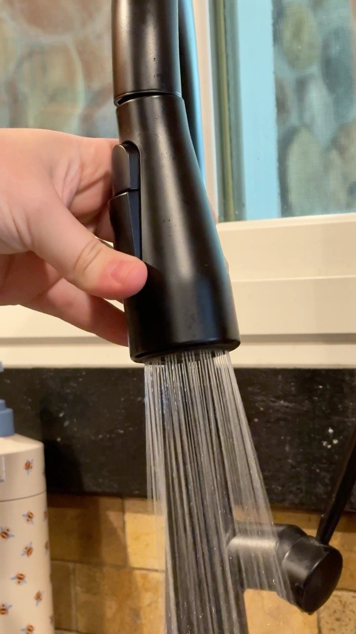 Matte black pull-down kitchen faucet spraying water while being held by a hand.
