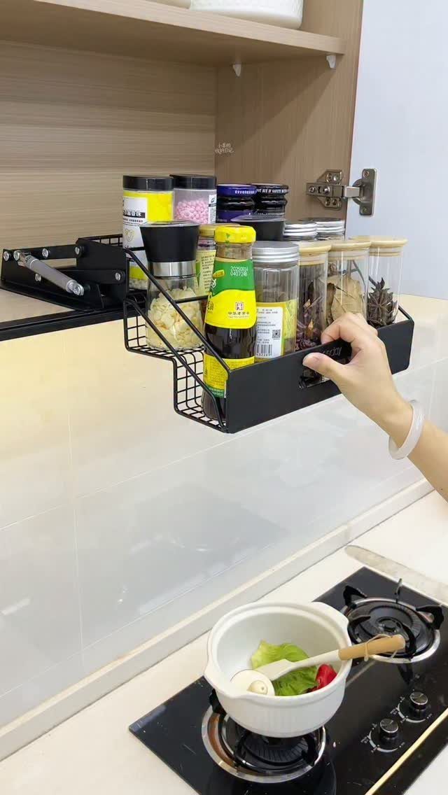 Black wire pull-out spice rack filled with jars inside a light wood cabinet.