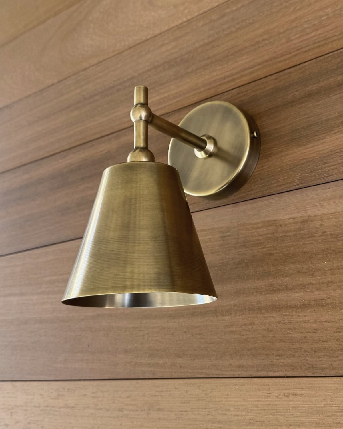 Brass wall sconce with a conical shade mounted on brown wooden planks.