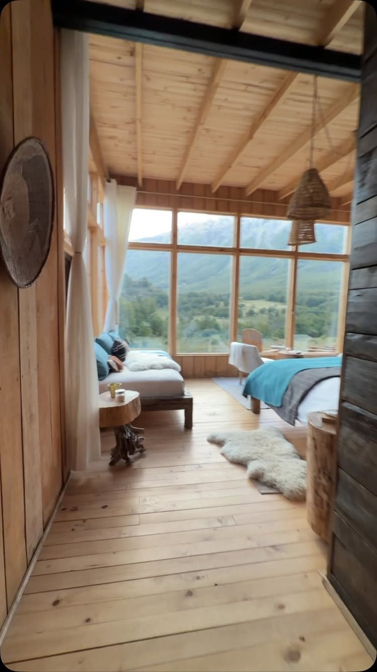 Sunlit rustic bedroom with light wood walls and large windows overlooking green mountains.