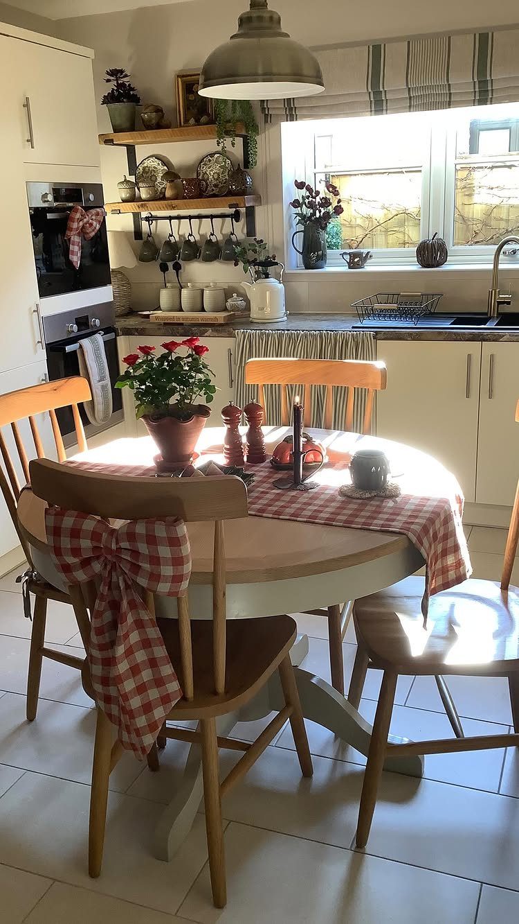 Round light-wood pedestal dining table with checkered runner and wooden chairs in a kitchen.