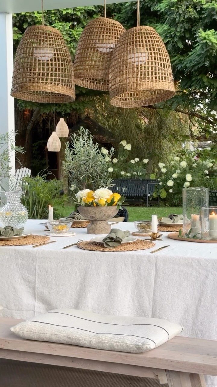 Outdoor dining table with white linen cloth, woven pendant lights, and garden backdrop