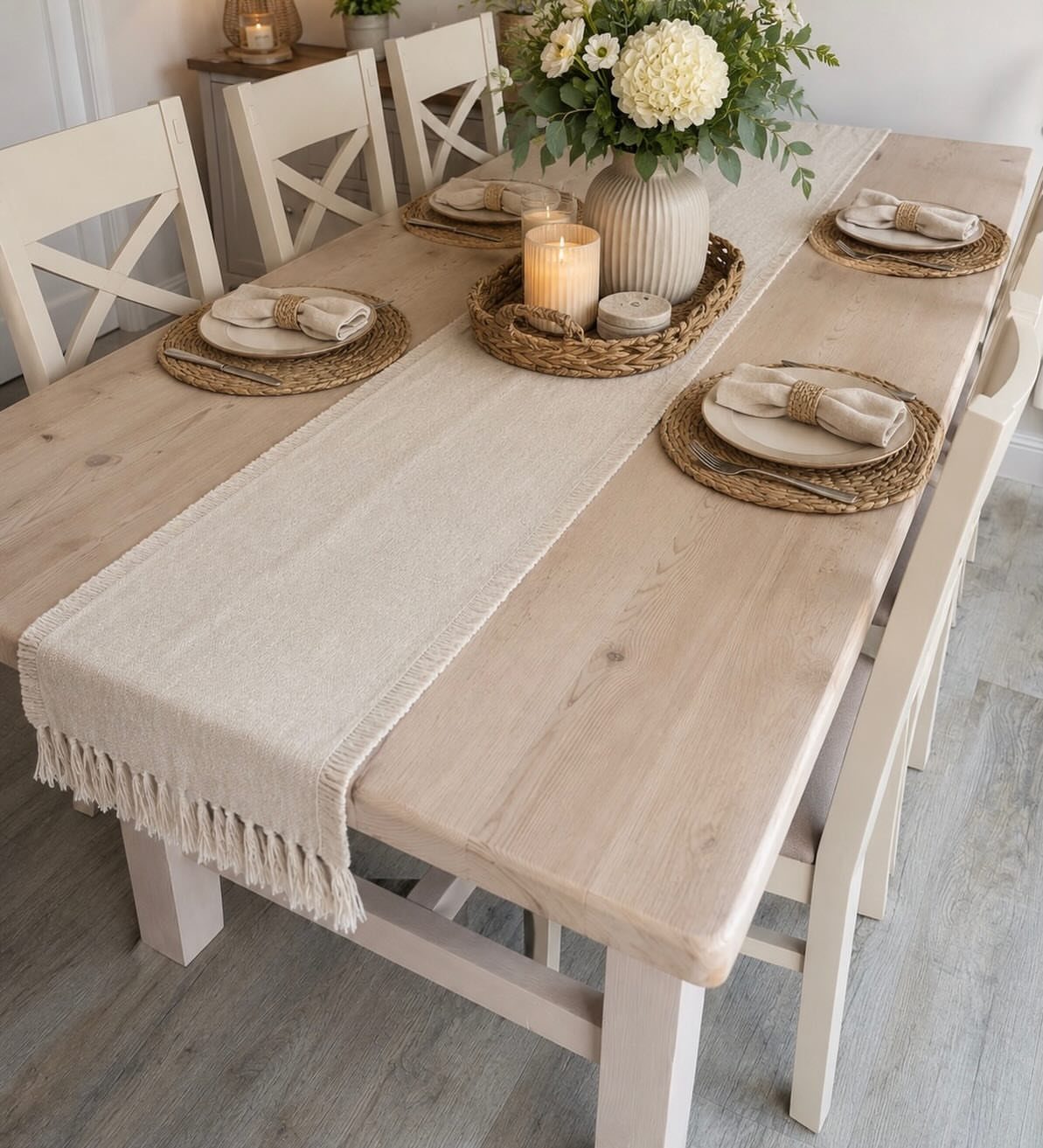 Light wood dining table with a beige fabric runner and white floral centerpiece.
