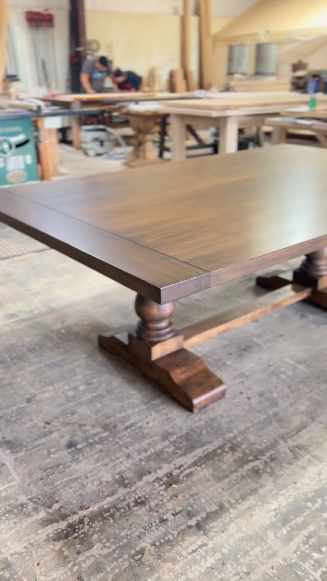 Dark brown wooden trestle dining table with turned legs in a woodworking shop.